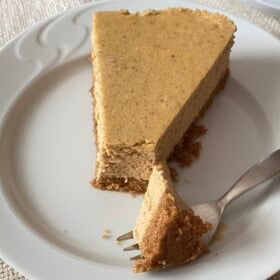 A slice of pumpkin cheesecake with a graham cracker crust on a plate with a fork cutting a bite.