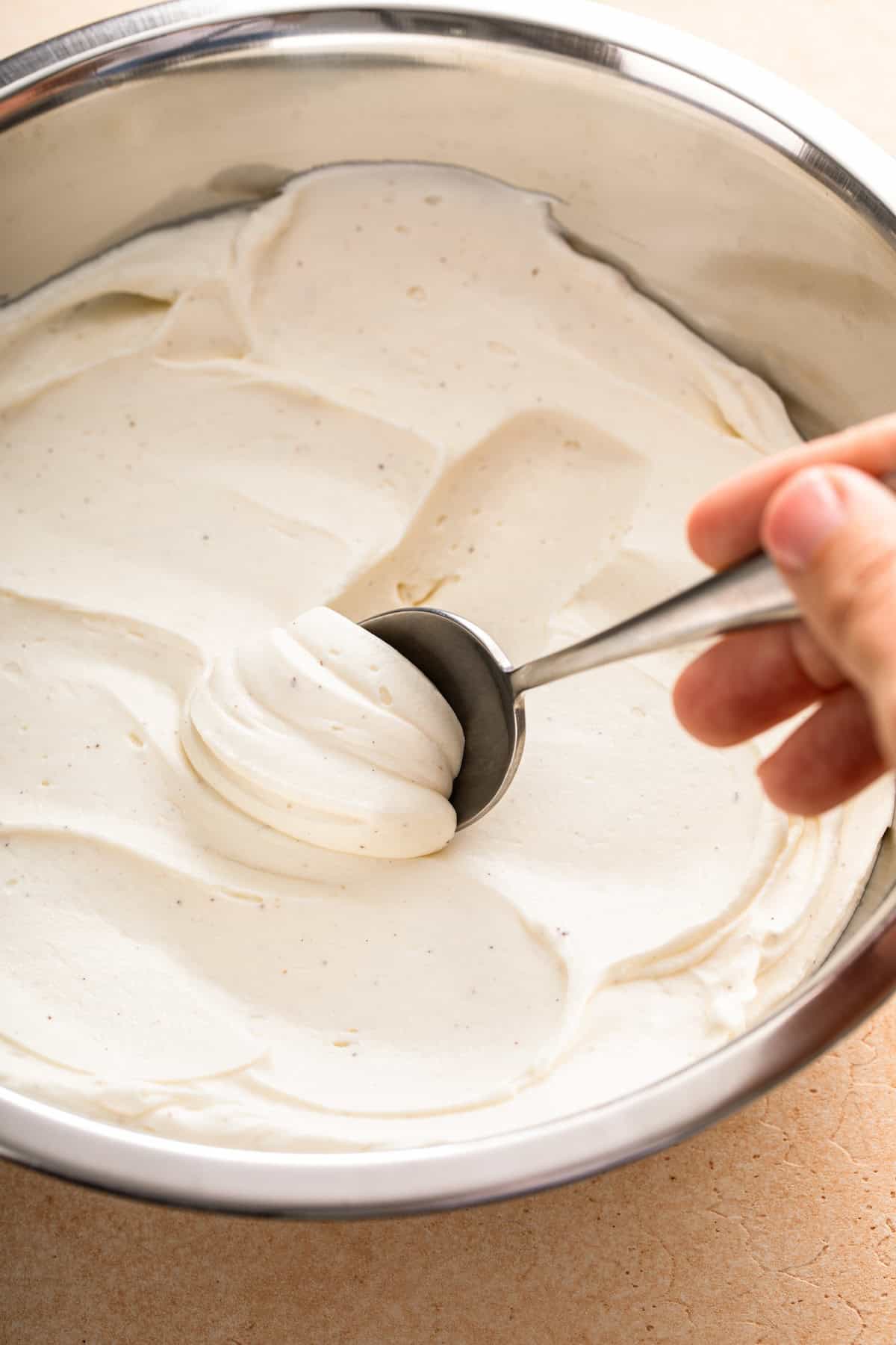 A spoon is scooping some whipped cream cheese frosting out of a large mixing bowl.