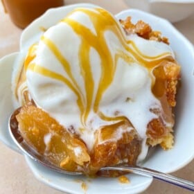 A serving of warm caramel apple dump cake in a bowl with ice cream on top and a spoon scooping up a bite.