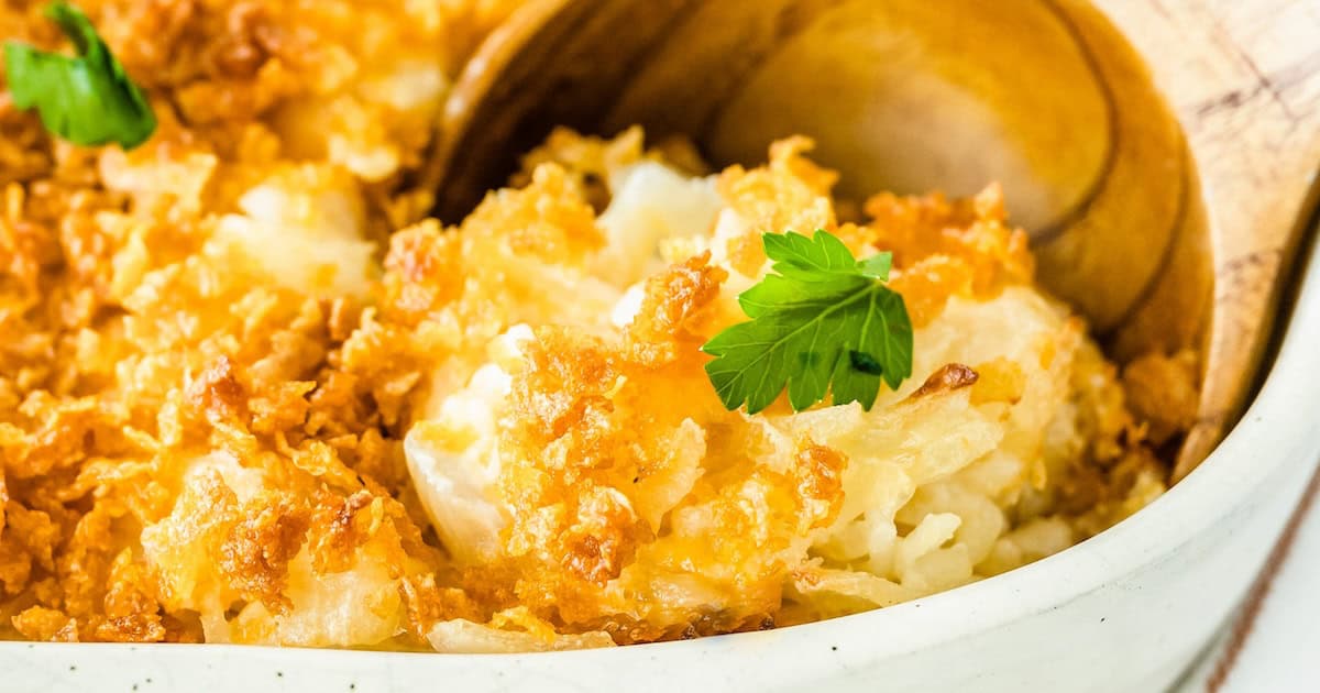 A wooden spoon scooping out a serving of cheesy potato casserole with cornflakes out of a baking dish.