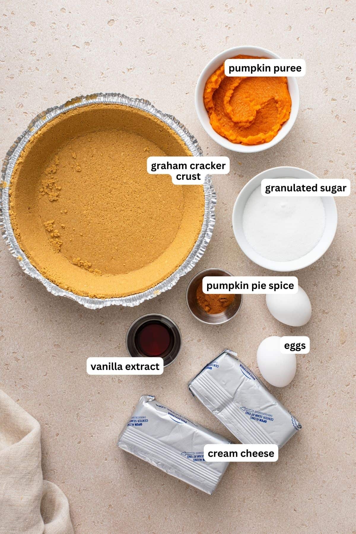 Ingredients for pumpkin pie cheesecake recipe in order from top to bottom: pumpkin puree, graham cracker crust, granulated sugar, cinnamon, vanilla extract, eggs, cream cheese.