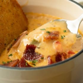 A spoon lifting a bite of beer and cheese soup out of a bowl with bacon and herbs on top.