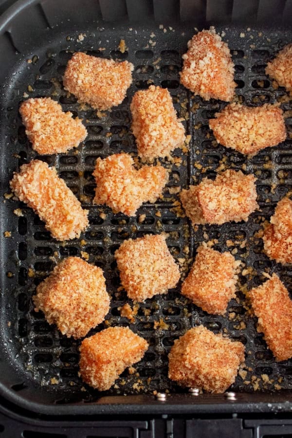 A layer of uncooked, seasoned salmon chunks are placed in the basket of an air fryer.