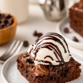 A scoop of vanilla ice cream on top of a homemade brownie is topped with a drizzle of chocolate sauce.