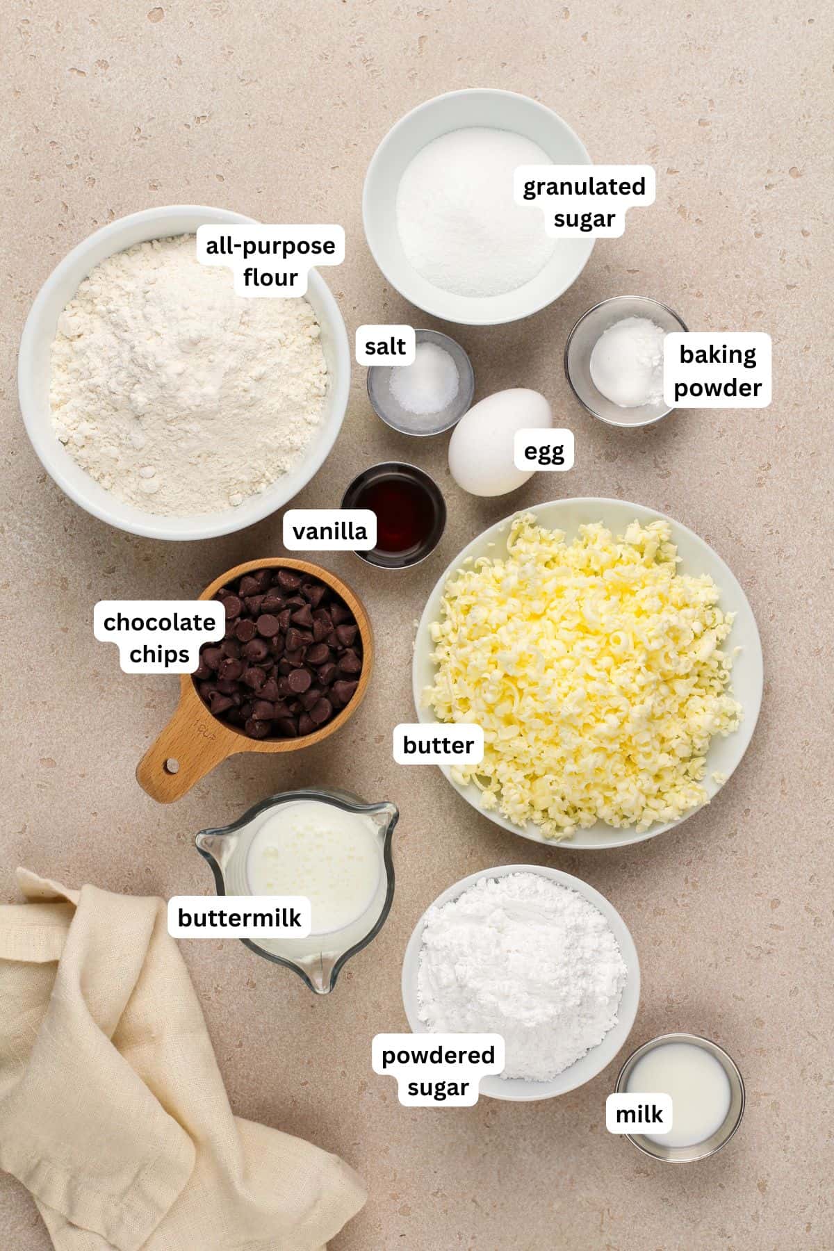 The ingredients for chocolate chip scone recipe in order from top to bottom: all purpose flour, granulated sugar, salt, egg, baking powder, vanilla, chocolate chips, butter, buttermilk, powdered sugar, milk.
