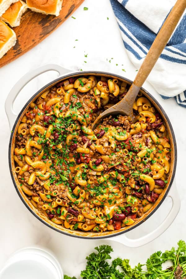 A cheesy, beefy skillet of goulash is being stirred with a large wooden spoon.