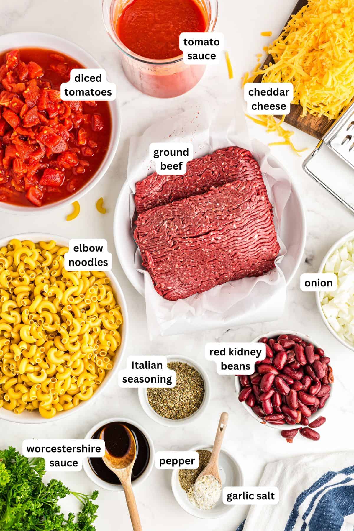 The ingredients for American goulash recipe in order from top to bottom: diced tomatoes, tomato sauce, cheddar cheese, ground beef, elbow noodles, onion, red kidney beans, Italian seasoning, Worcestershire sauce, pepper, garlic salt.