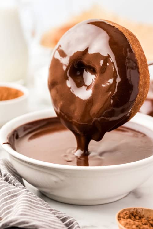 A donut is being dipped into a bowl filled with glossy chocolate glaze.