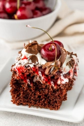 A slice of easy Black Forest cake on a white plate, topped with whipped cream, chocolate shavings, and two real cherries on top. A bowl of cherries is blurred in the background on a light-colored surface.
