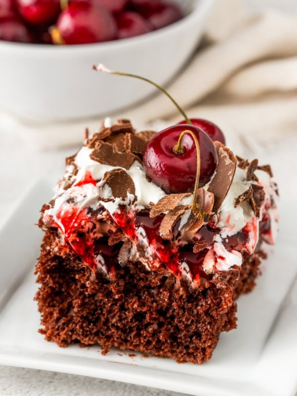 A slice of easy Black Forest cake on a white plate, topped with whipped cream, chocolate shavings, and two real cherries on top. A bowl of cherries is blurred in the background on a light-colored surface.