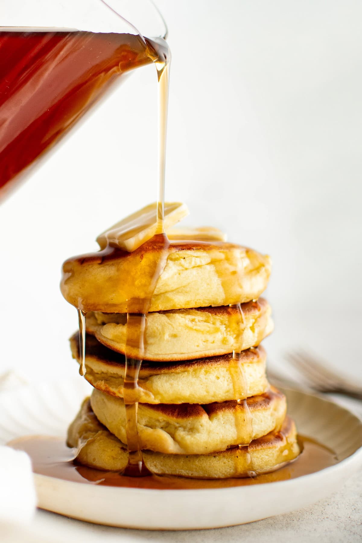 A stack of fluffy Japanese pancakes topped with butter is on a plate, with syrup being poured over them, cascading down the sides. A fork is partially visible in the background.