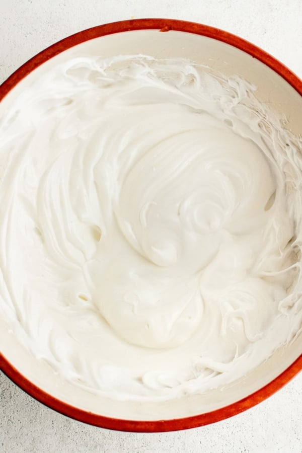 A red-rimmed bowl filled with smooth, fluffy white whipped egg whites, showcasing its soft peaks and creamy texture, placed on a light-colored surface.