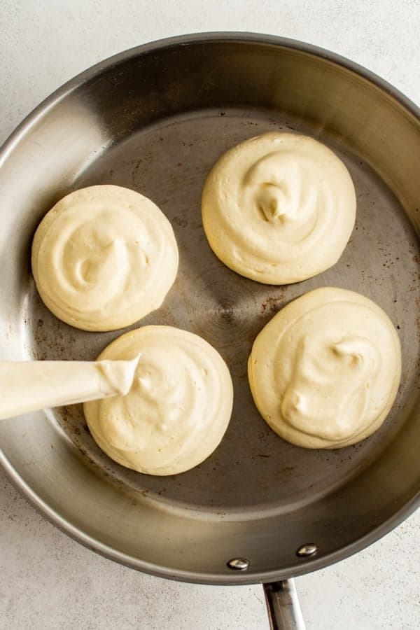 A stainless steel pan containing four dollops of pancake batter, with a piping bag adding another swirl. The batter is smooth and light, ready to be cooked. The background is a light gray surface.