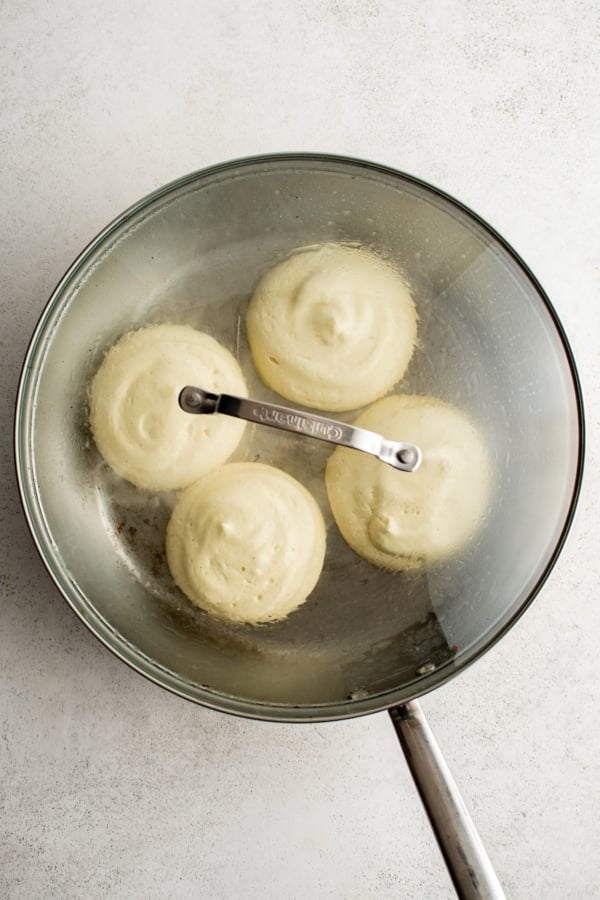 A frying pan with a glass lid containing four fluffy, round pancakes cooking. The pancakes are steaming inside the covered pan.