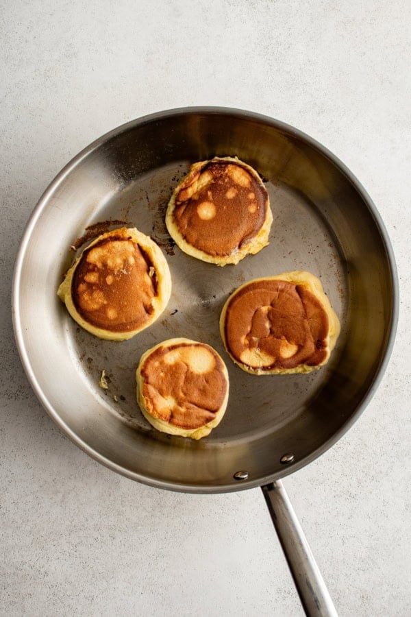 A metal skillet containing four golden-brown, fluffy pancakes. The pancakes have a slightly uneven surface and are arranged in a circular pattern. The skillet rests on a light, textured surface.