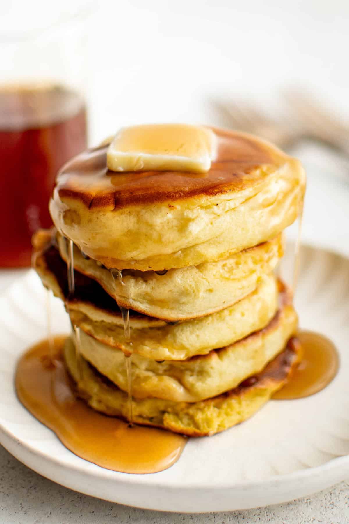 A stack of fluffy soufflé pancakes topped with a pat of butter and drizzled with syrup sits on a plate. A glass bottle of syrup is visible in the background.