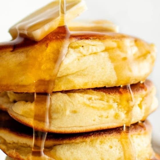 A stack of three fluffy soufflé pancakes topped with butter and maple syrup dripping down the sides. The background is softly blurred, emphasizing the golden-brown pancakes.