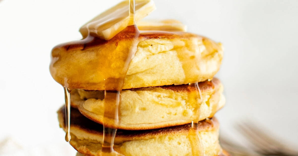 A stack of three fluffy soufflé pancakes topped with butter and maple syrup dripping down the sides. The background is softly blurred, emphasizing the golden-brown pancakes.