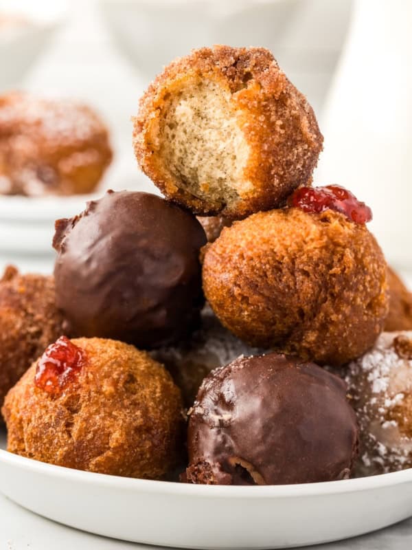 A plate of assorted donut holes, including chocolate-covered, powdered sugar, and jelly-filled varieties. The top cinnamon sugar donut hole has a bite taken out, revealing its fluffy interior.