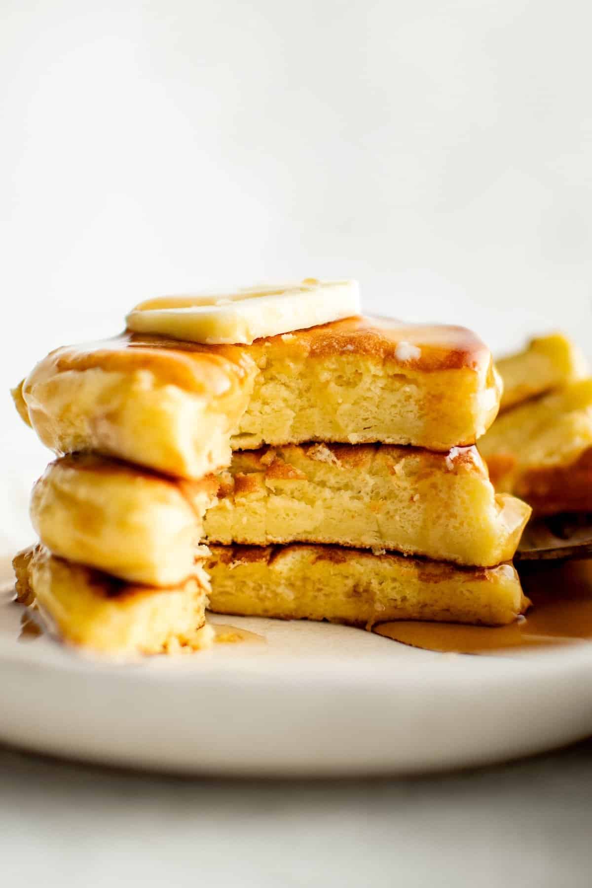 A stack of three fluffy Japanese soufflé pancakes topped with a slice of butter and drizzled with syrup, on a white plate. A bite is taken out of the pancakes, revealing their soft, light and airy interior.