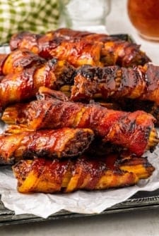 Close-up of a tray filled with bacon-wrapped stuffed shotgun shells, glazed with a shiny barbecue sauce. They are neatly arranged on crinkled parchment paper, with a checkered cloth napkin visible in the background.