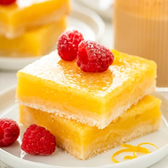 Two thick lemon bars with a shortbread crust stacked on a white plate, topped with fresh raspberries. A spiral of lemon zest decorates the plate.