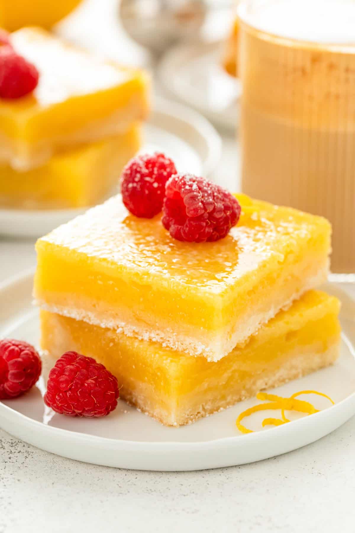 Two thick lemon bars with a shortbread crust stacked on a white plate, topped with fresh raspberries. A spiral of lemon zest decorates the plate.