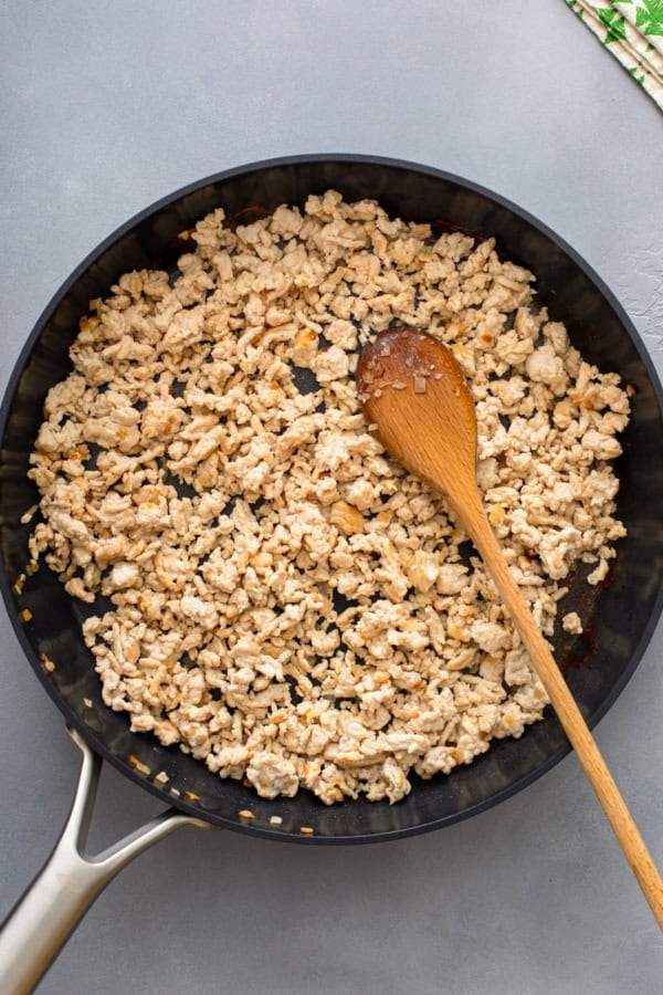 A skillet filled with cooked ground chicken, being stirred with a wooden spoon, sits on a gray countertop. A small portion of a green patterned cloth is visible in the top right corner.