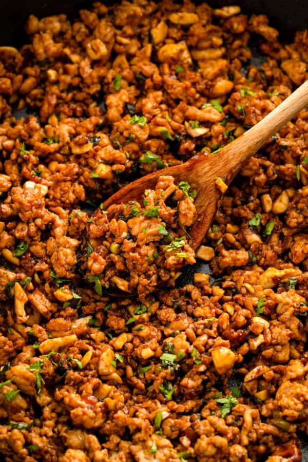 Close-up of a wooden spoon stirring a skillet of cooked ground chicken mixed with a savory brown sauce, chopped vegetables, and garnished with fresh cilantro.