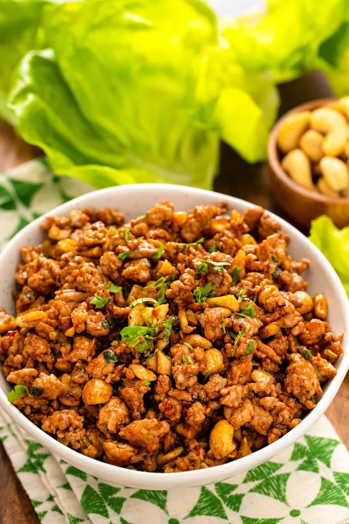 A white bowl filled with seasoned ground chicken filling garnished with cilantro, sitting on a patterned cloth. In the background, there are fresh lettuce leaves and a small bowl of cashews.