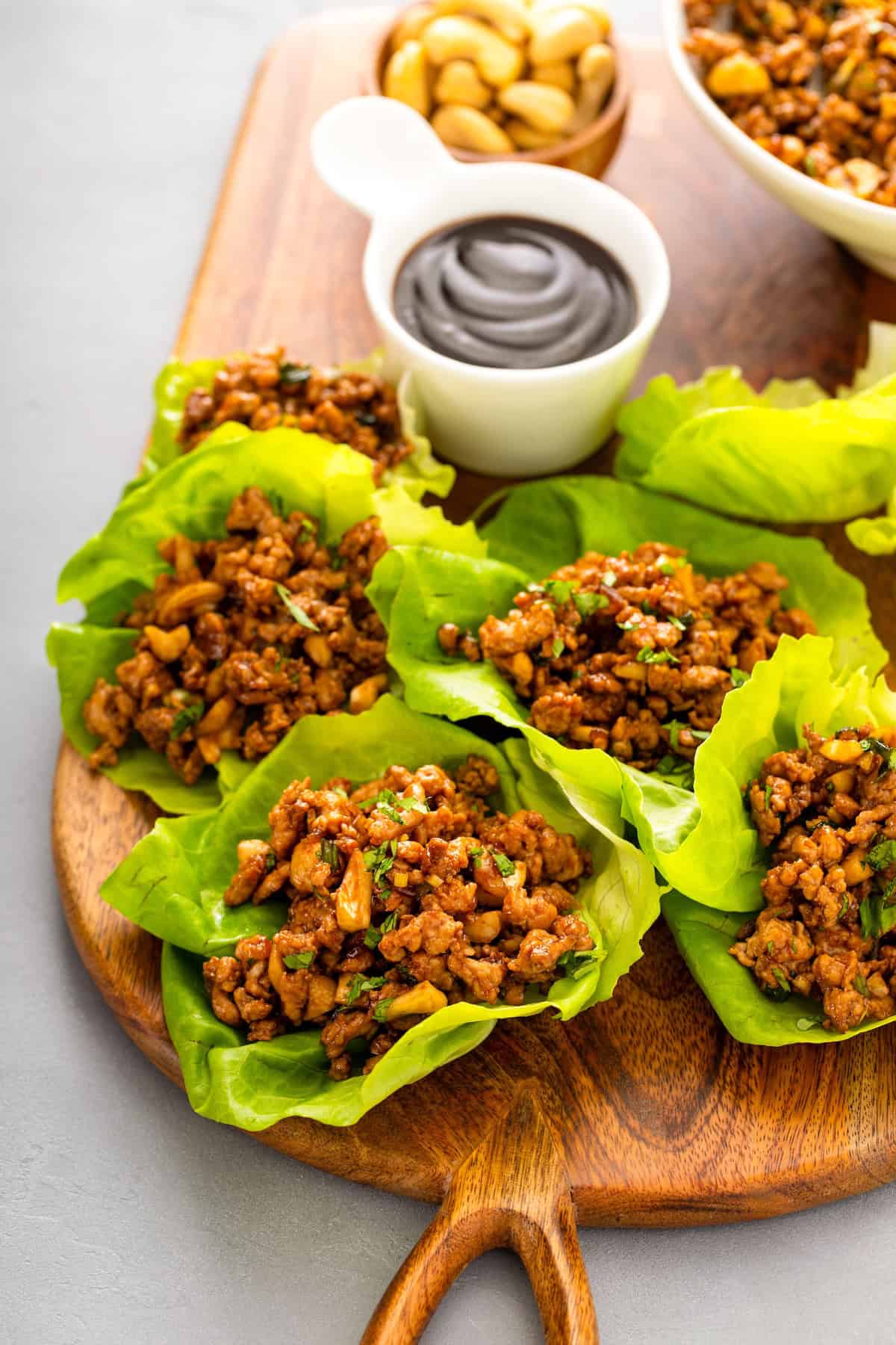 A wooden board with several chicken lettuce wraps filled with a savory ground chicken mixture, garnished with cilantro. A small bowl of dark dipping sauce and a bowl of cashews sit nearby.