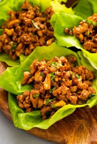 Three lettuce leaves filled with a savory ground chicken mixture with chopped cashews and fresh cilantro on top, served on a wooden plate.