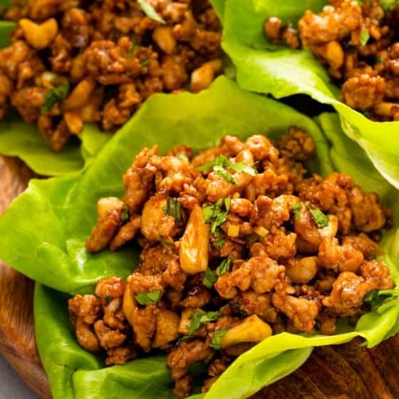 Three lettuce leaves filled with a savory ground chicken mixture with chopped cashews and fresh cilantro on top, served on a wooden plate.
