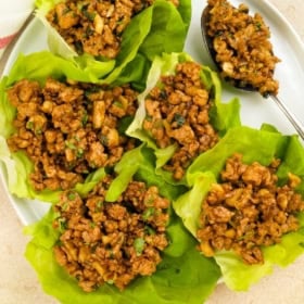 Six lettuce wraps filled with a cooked, seasoned ground chicken mixture are arranged on a white plate, with a spoonful of the same filling placed on the side. The filling appears to contain herbs and diced vegetables.