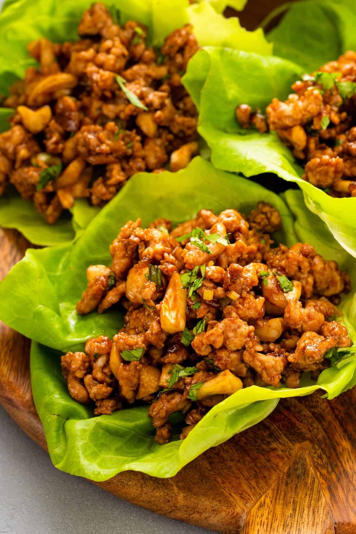 Ready in 20 minutes, these P.F. Chang’s inspired Chicken Lettuce Wraps are loaded with fresh garlic, ginger, lemongrass, and a touch of heat. Three lettuce leaves filled with a savory ground chicken mixture with chopped cashews and fresh cilantro on top, served on a wooden plate.