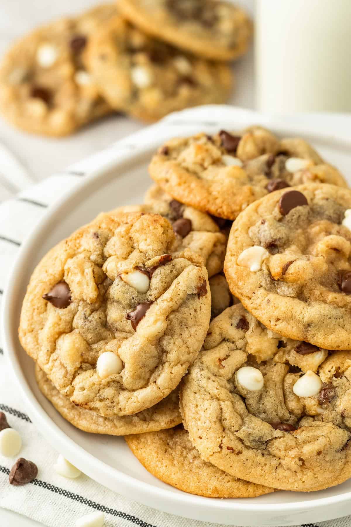A plate of freshly baked chocolate chip walnut cookies with both white and milk chocolate chips, stacked together on a white plate, with more cookies and a glass of milk blurred in the background.