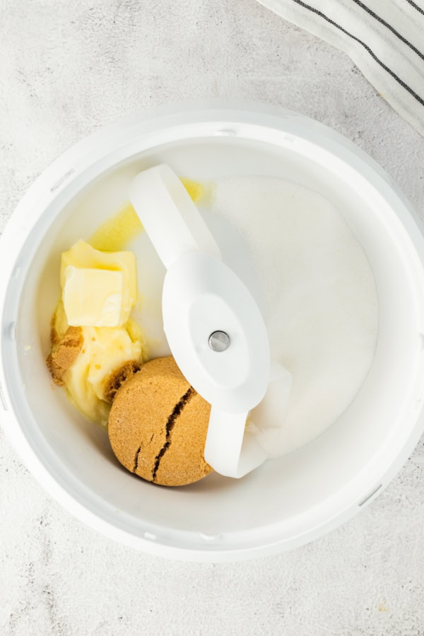 A mixing bowl with white sugar, brown sugar, and chunks of butter, ready to be mixed. The bowl sits on a light countertop next to a striped cloth.