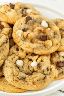 A close-up of a plate piled with freshly baked chocolate chip walnut cookies, featuring both white and dark chocolate chips, on a white background.