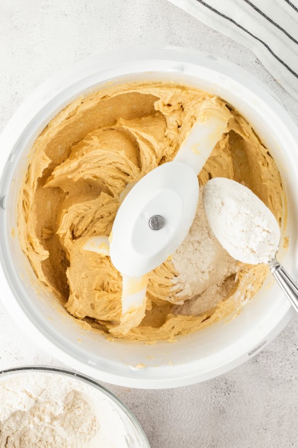 Overhead view of cookie dough being mixed in a white electric mixer; a spoon holds flour above the bowl, ready to be added. A bowl of flour and a striped cloth are partially visible nearby.