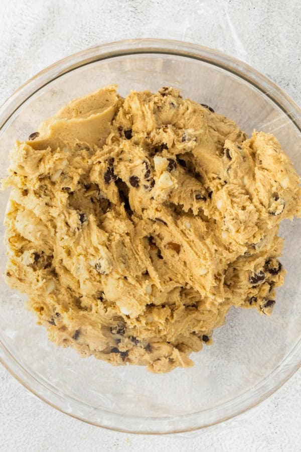 A glass bowl filled with cookie dough sits on a light-colored textured surface. The dough appears creamy and thick, with visible chocolate chips and walnuts mixed throughout.