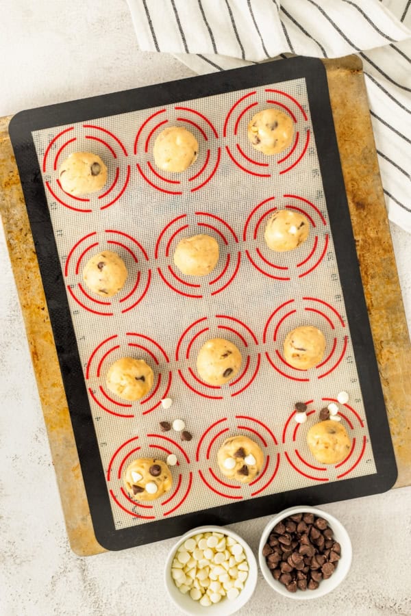 A baking sheet lined with a silicone mat holds 12 unbaked cookie dough balls, some with chocolate chips pressed into the top. Nearby are three small bowls filled with white chocolate chips and regular chocolate chips.