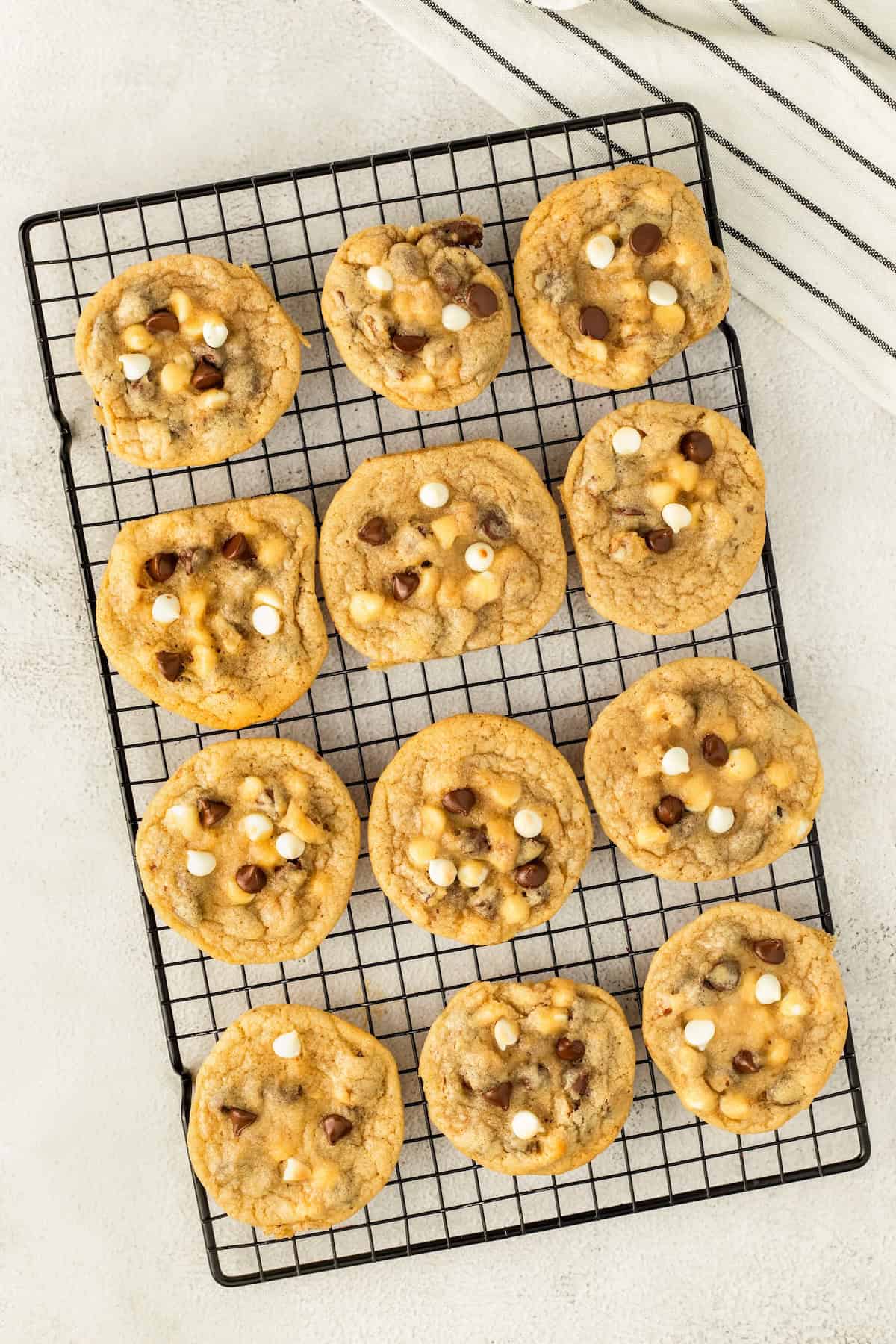 Twelve freshly baked chocolate chip and walnut cookies with white and milk chocolate chips are arranged on a black cooling rack, set on a light-colored surface. A white towel with black stripes is partially visible in the corner.