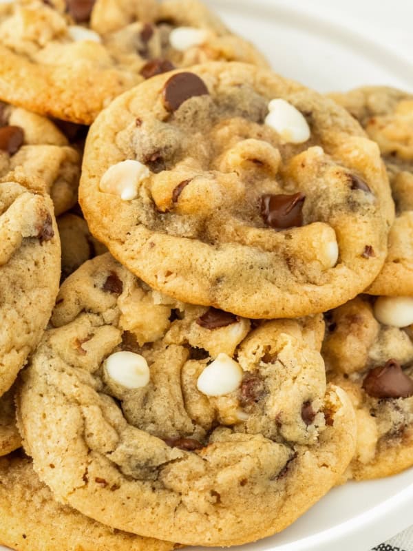 A close-up of a plate piled with freshly baked chocolate chip walnut cookies, featuring both white and dark chocolate chips, on a white background.