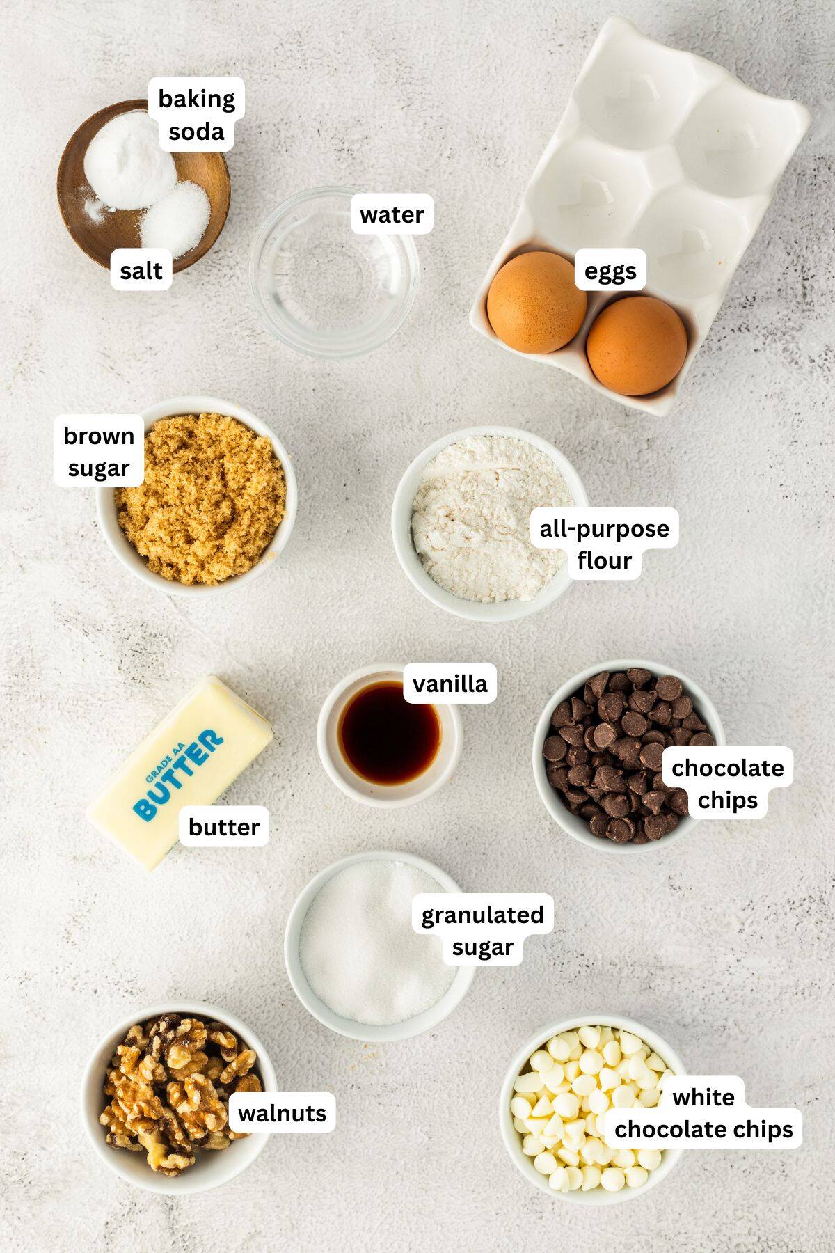 Flat lay of chocolate chip walnut cookie recipe ingredients in bowls on a light surface, including eggs, butter, brown sugar, granulated sugar, salt, baking soda, flour, vanilla, walnuts, chocolate chips, white chocolate chips, and water.