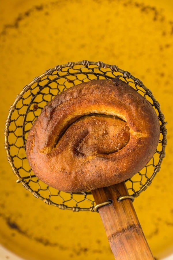 A golden-brown fried bun is held above a pot of yellow oil using a wire skimmer with a wooden handle.