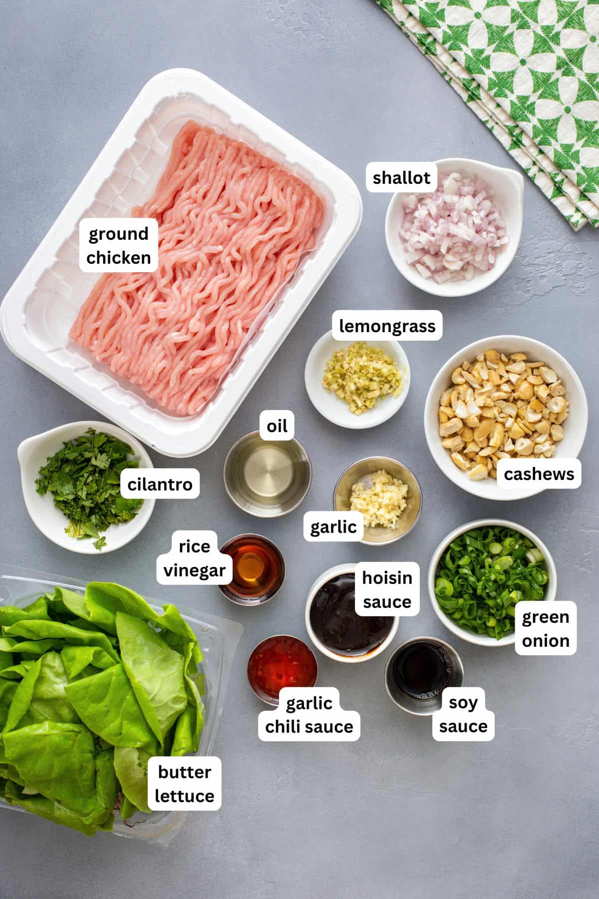 Ingredients for chicken lettuce wraps recipe arranged on a gray surface, including ground chicken, minced shallot, lemongrass, cashews, cilantro, oil, green onion, garlic, rice vinegar, hoisin sauce, soy sauce, garlic chili sauce, and butter lettuce.