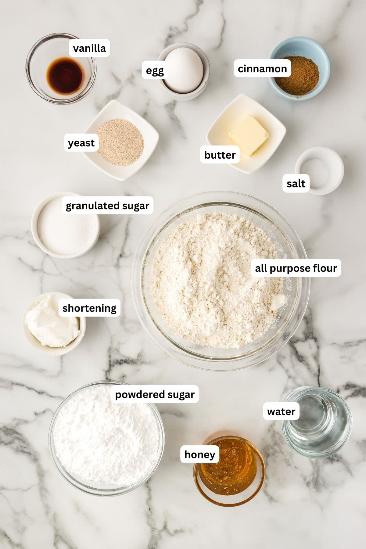 Ingredients for homemade honey buns recipe are arranged on a marble surface, each labeled: vanilla, egg, yeast, granulated sugar, shortening, powdered sugar, all-purpose flour, butter, cinnamon, salt, water, and honey.