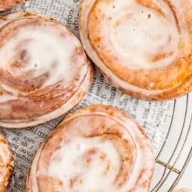 Glazed homemade honey buns on a wire rack with a newspaper underneath.