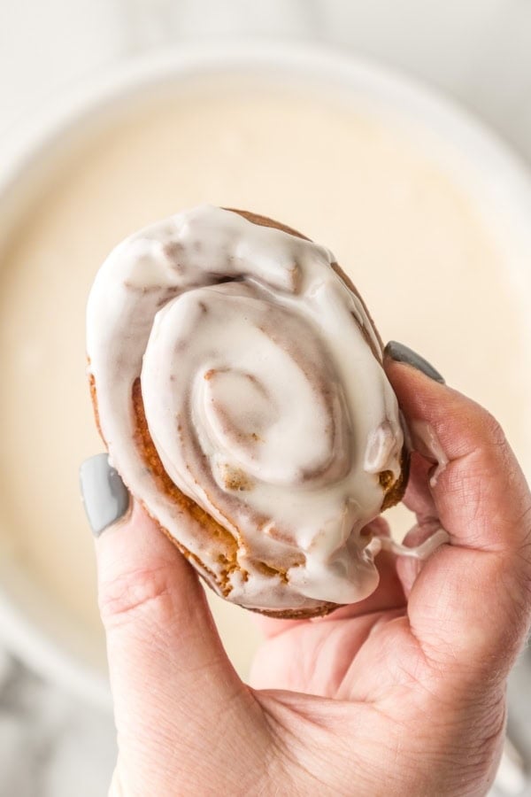 A hand holding a frosted honeybun close to the camera, with a bowl of icing blurred in the background.