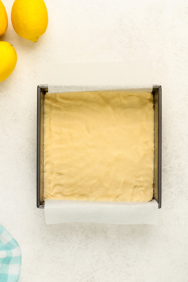 A baking dish lined with parchment paper contains unbaked shortbread dough spread evenly. Three lemons are visible in the top left corner, and part of a blue checkered cloth is in the bottom left corner.